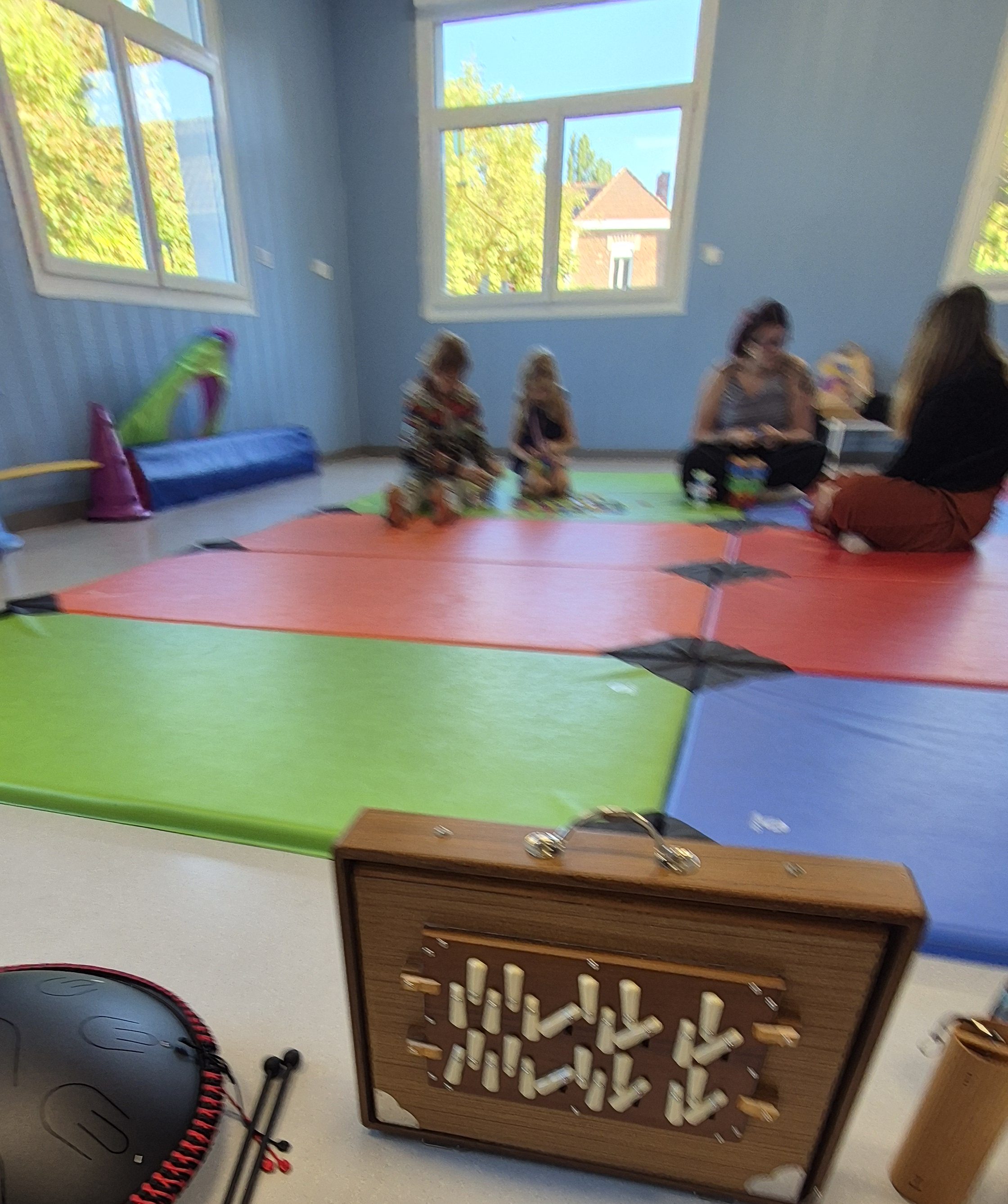 Une salle de jeu colorée avec des tapis orange, vert et bleu. Des enfants jouent ensemble au sol pendant qu'un adulte les surveille. Des instruments de musique sont visibles à l'avant-plan.