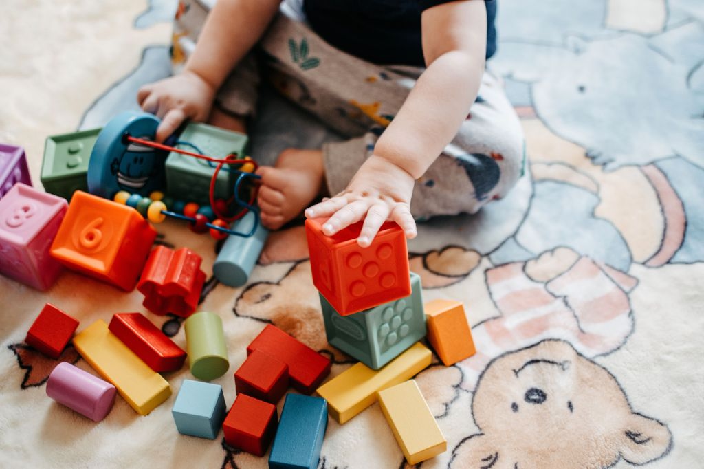 Un enfant joue avec des blocs de construction colorés sur un tapis illustré, utilisant une main pour empiler un bloc rouge sur une pile.
