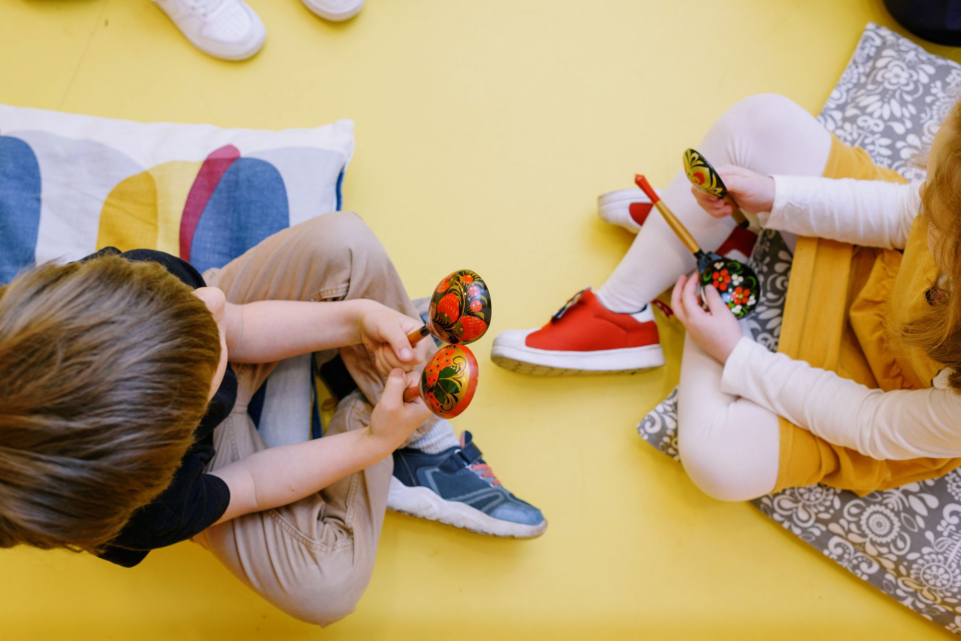 Enfants assis sur un sol jaune, tenant des œufs en bois peints colorés. L'un porte un t-shirt noir et des pantalons clairs, tandis que l'autre est en robe jaune avec des collants blancs et des chaussures rouges.