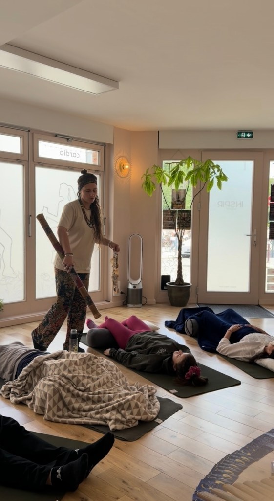 Une praticienne guide un groupe de personnes allongées sur des tapis dans un espace apaisant, avec des plantes en pot et une lumière douce.