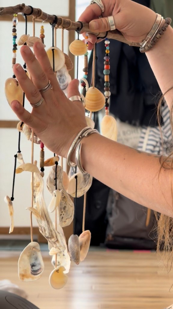 Une main tenant un mobile fait de coquillages et de perles colorées, suspendu à une tige en bois.
